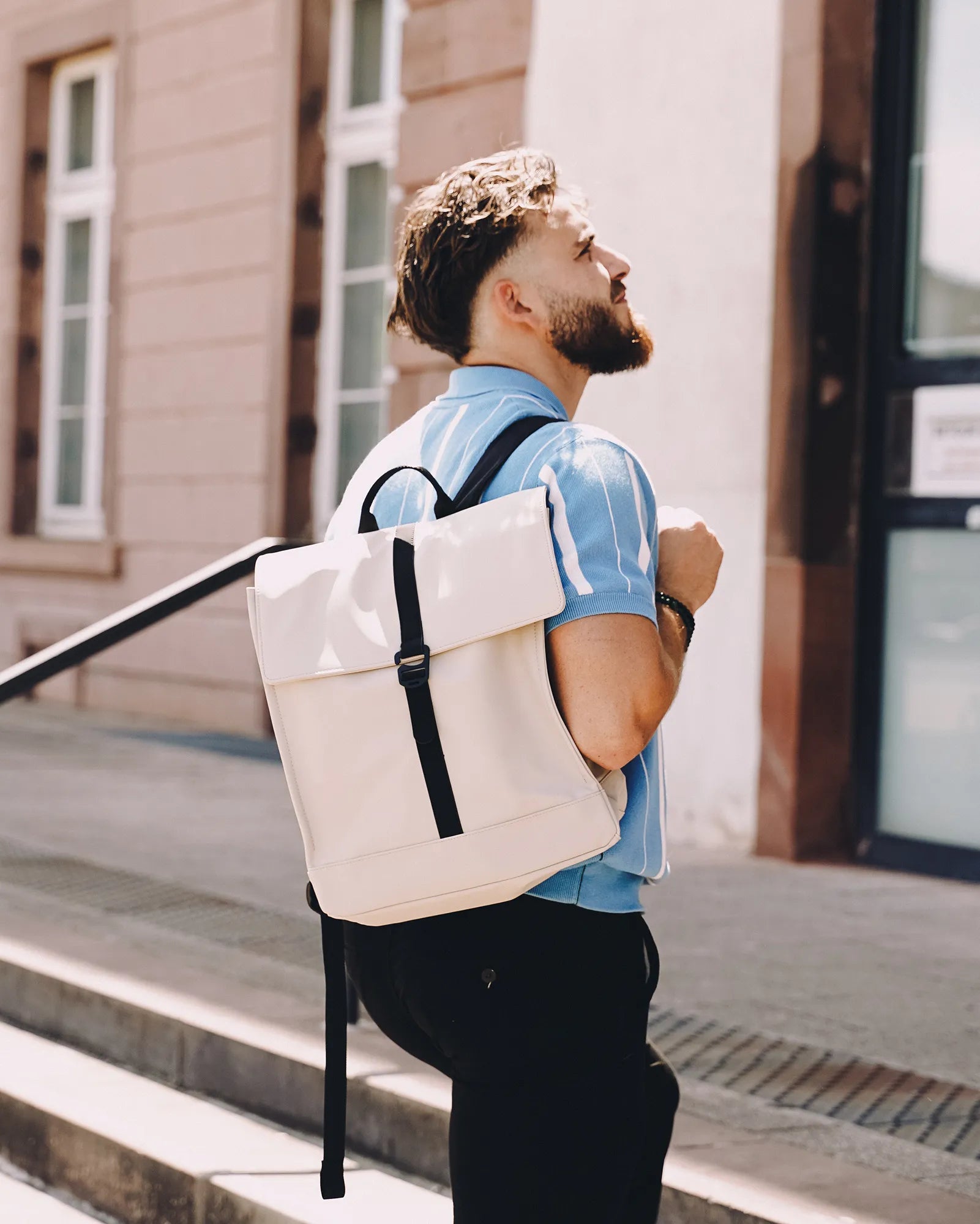 Homme portant un sac à dos blanc à la mode et regardant vers le haut à l'extérieur sur un trottoir ensoleillé.