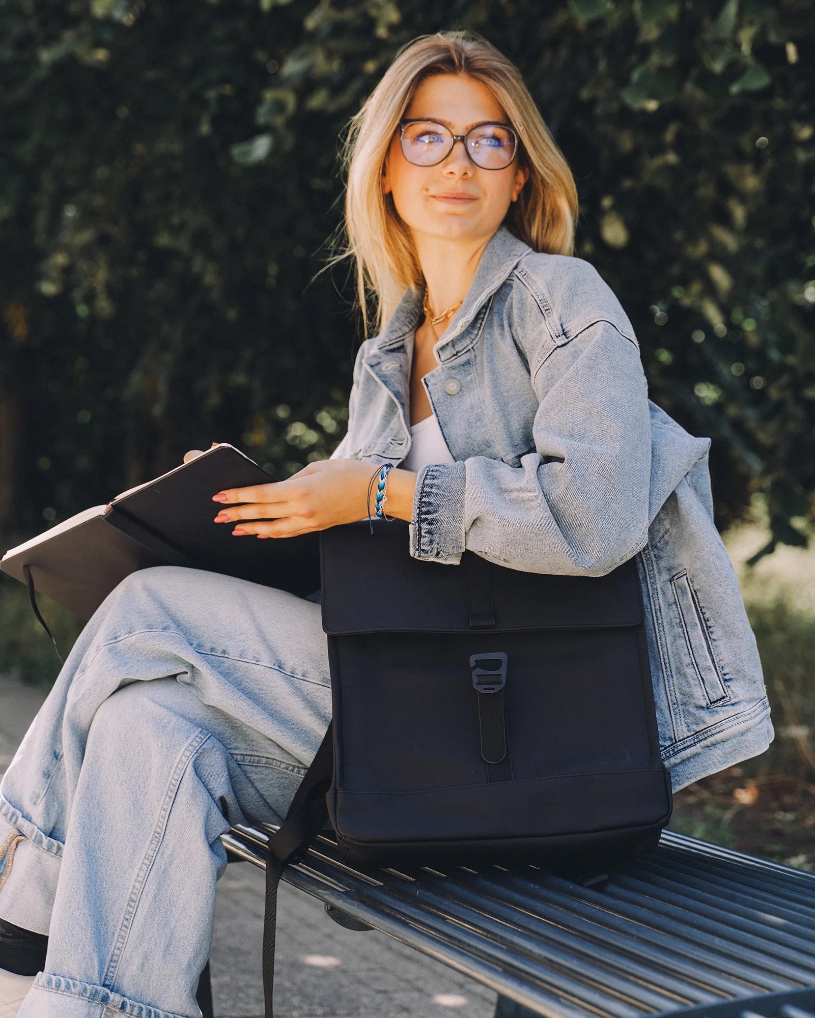 Jeune femme blonde portant une veste en jean et des lunettes, assise sur un banc en extérieur avec un carnet ouvert et un sac à dos noir.