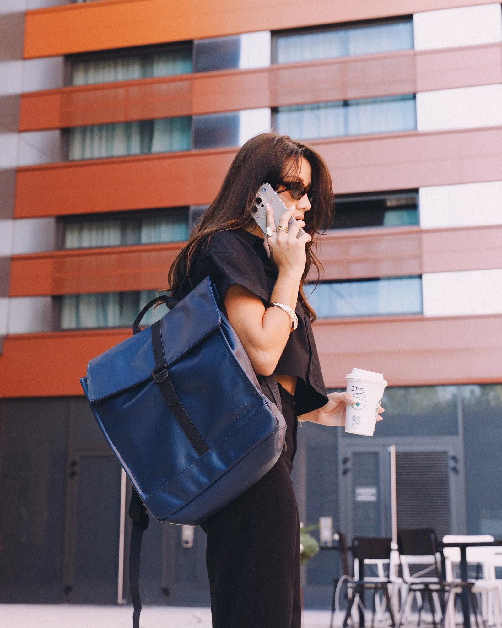 Femme portant un sac à dos bleu, tenant un café à emporter et parlant au téléphone devant un bâtiment moderne.