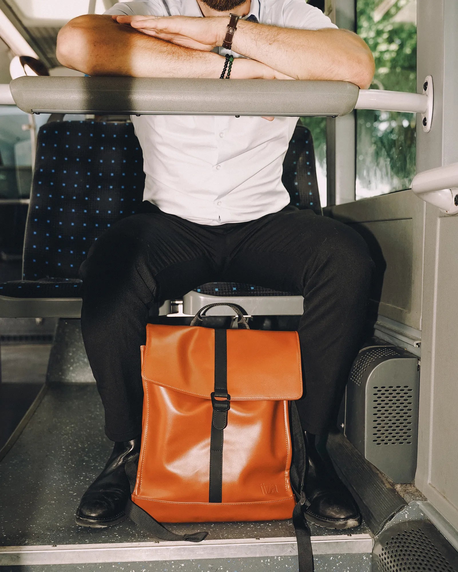 Homme assis dans un bus portant une chemise blanche et un pantalon noir avec un sac à dos cuir marron entre ses jambes.