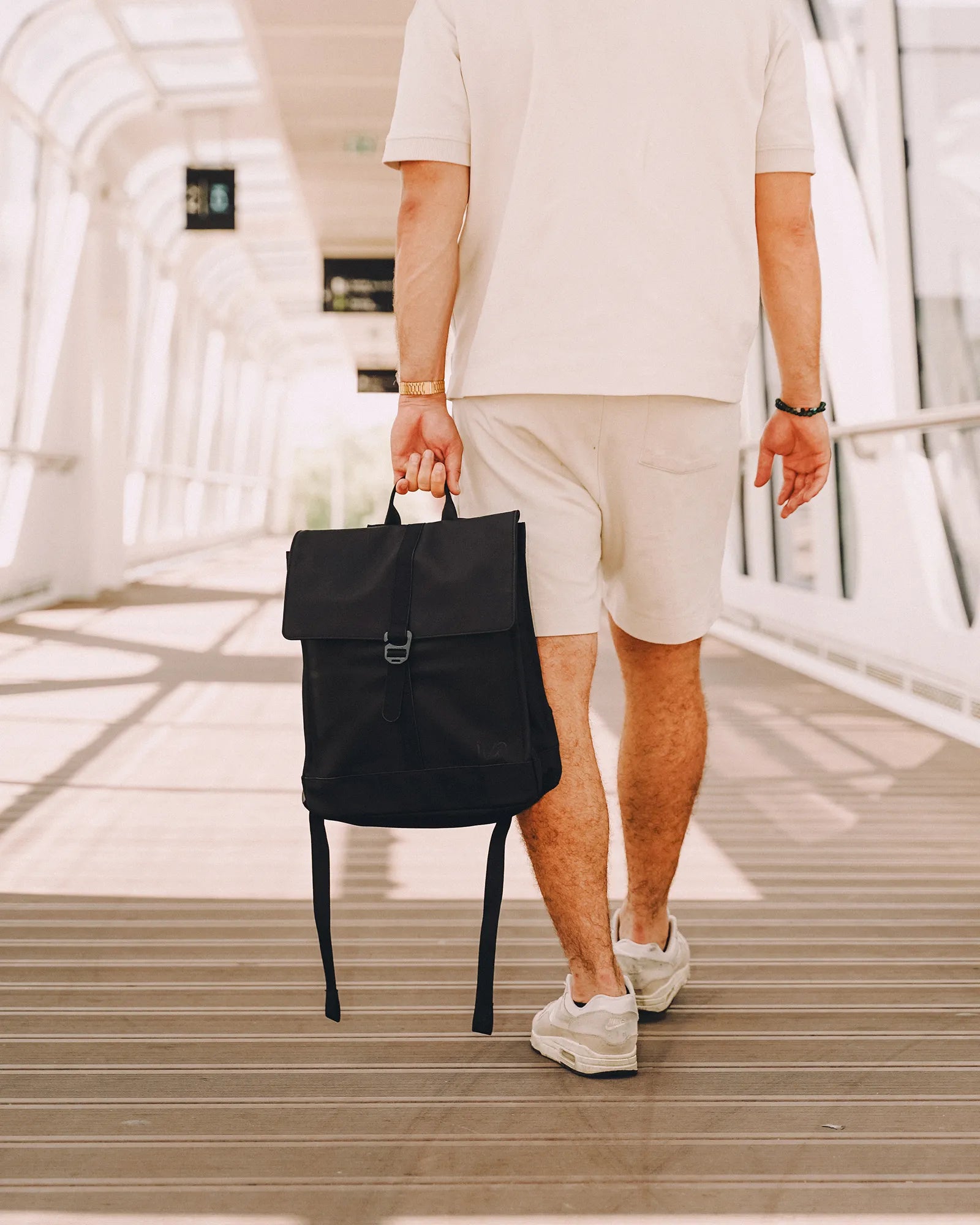 Homme marchant dans un passage lumineux portant un sac à dos noir à la main, vêtu d'un short et d'un polo blancs et de baskets blanches.
