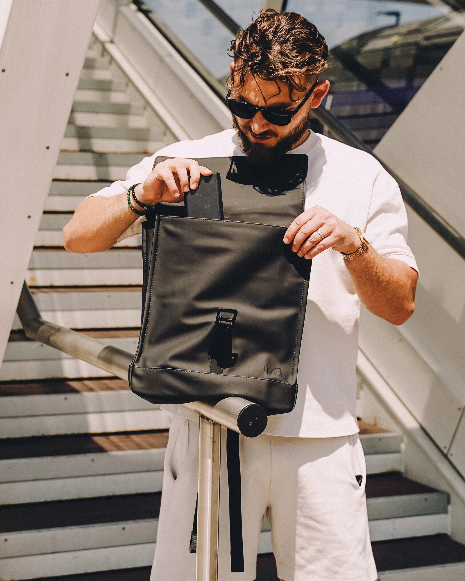 Un homme avec des lunettes de soleil range un téléphone portable dans un sac à dos noir devant un escalier extérieur.