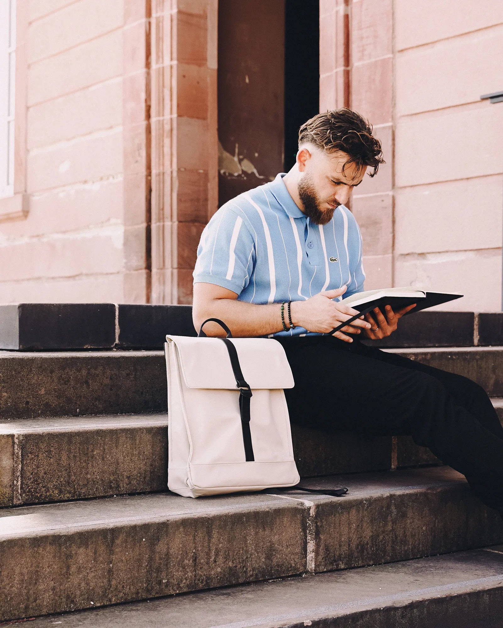 Un homme assis sur des marches en pierre lisant un livre, avec un sac à dos beige posé à côté de lui.
