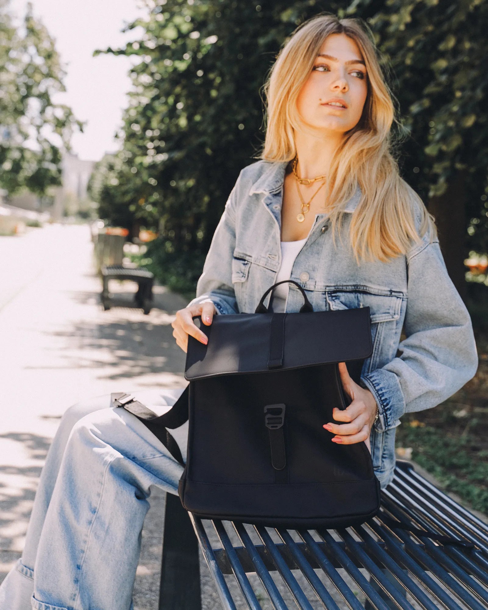 Jeune femme blonde en veste en jean assise sur un banc avec un sac à dos noir.