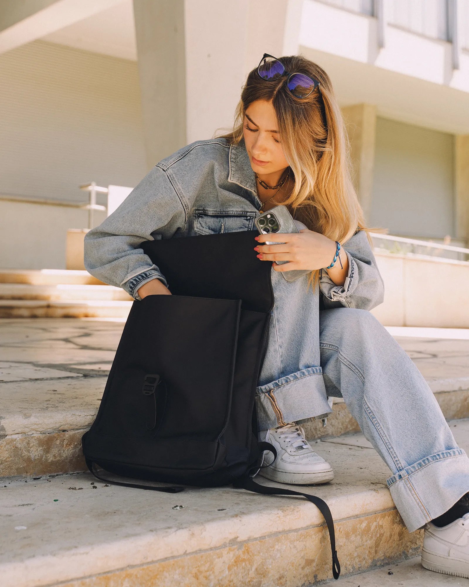 Jeune femme assise sur des marches en jean clair, rangeant un téléphone dans un sac à dos noir.