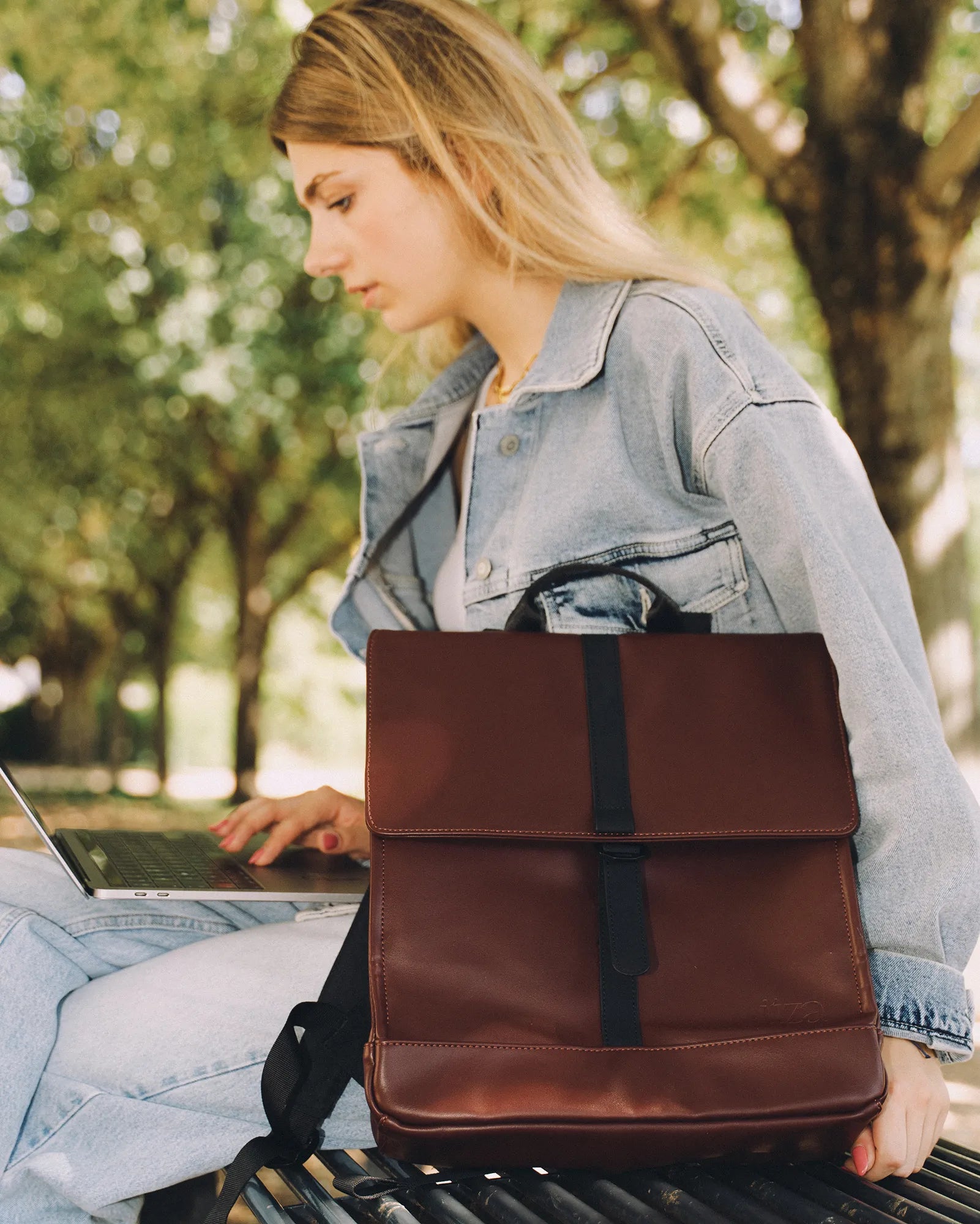 Jeune femme portant une veste en jean et un jean clair assise sur un banc avec un ordinateur portable et un sac à dos en cuir marron.