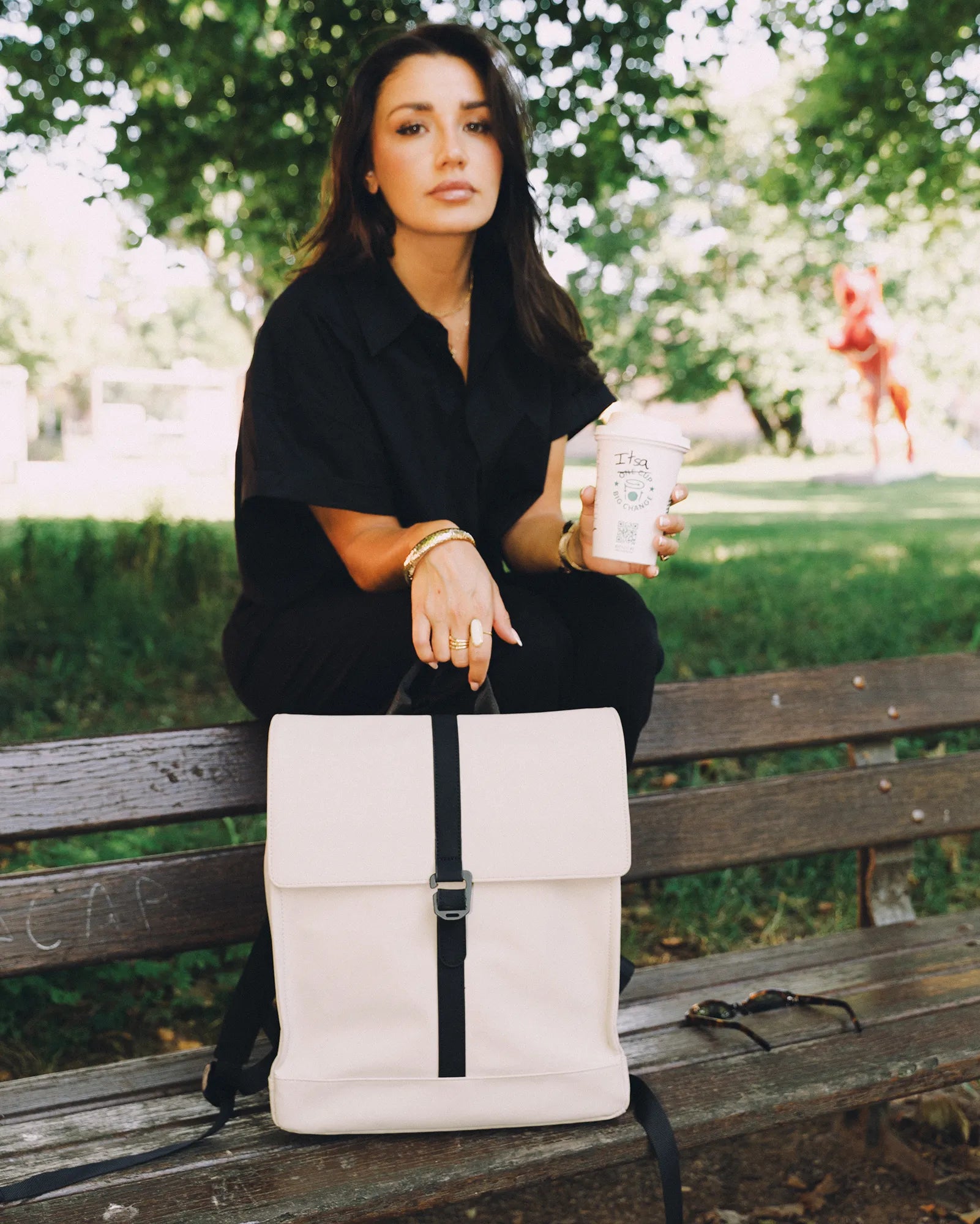 Femme assise sur un banc dans un parc, tenant un gobelet à café réutilisable et posant une main sur un sac à dos beige.