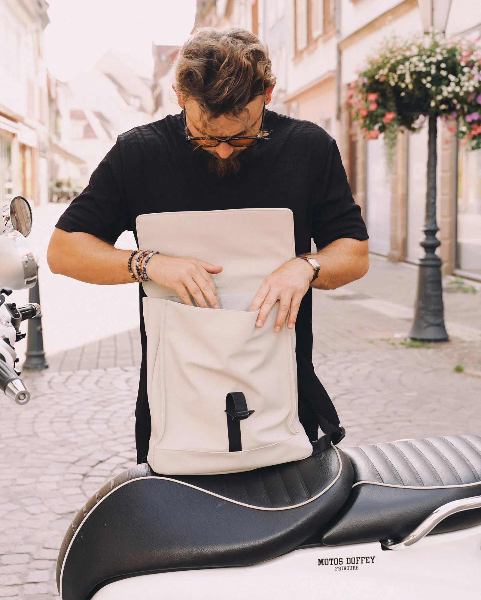 Un homme avec des lunettes cherche dans un sac à dos beige posé sur la selle d'une moto blanche garée sur une rue pavée.