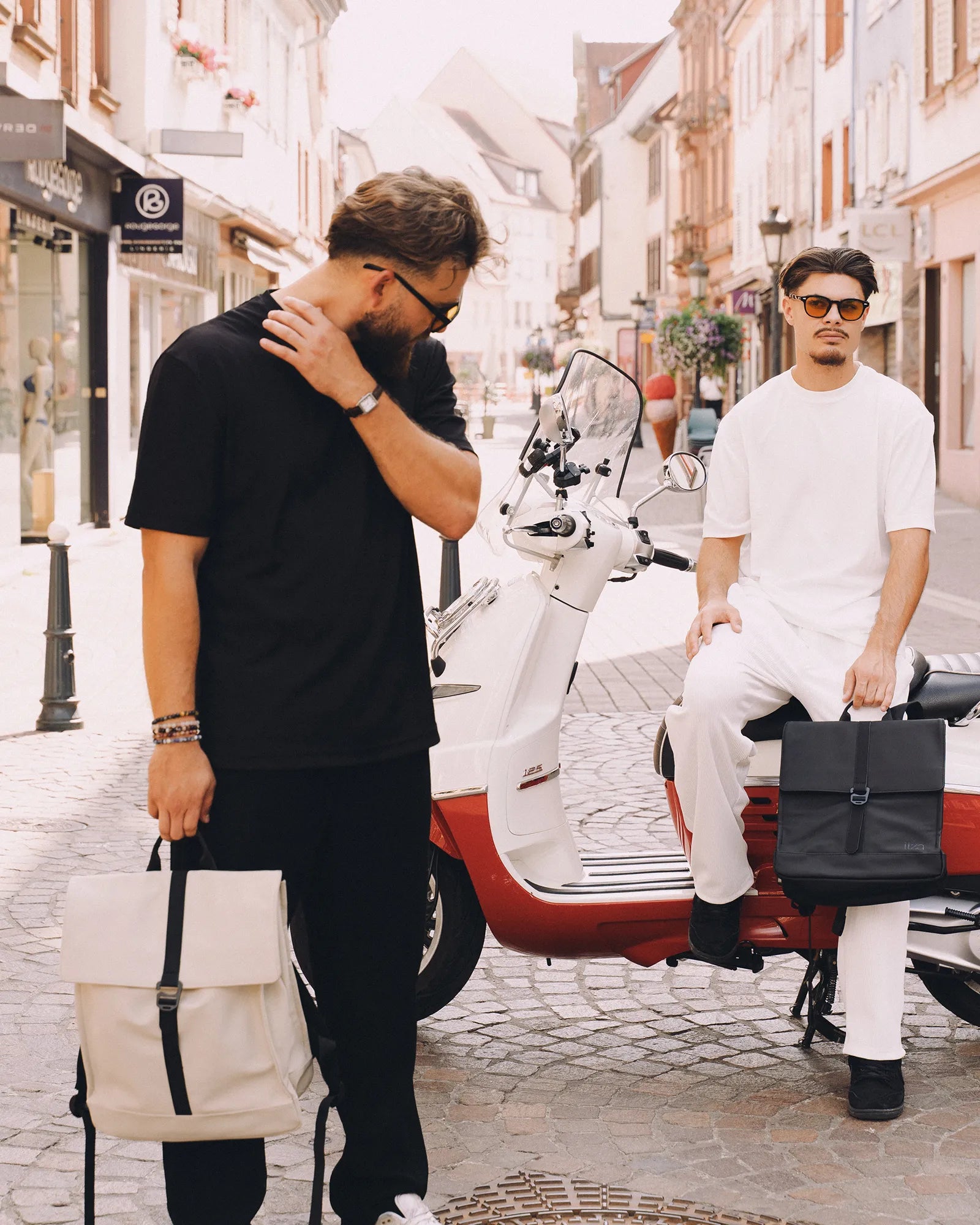 Deux hommes élégants portant des sacs à dos, l'un debout en noir tenant un sac crème et l'autre assis sur un scooter blanc et rouge avec un sac noir dans une rue pavée.