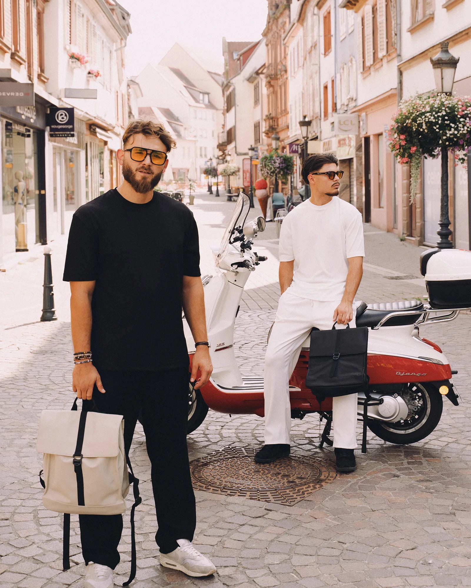 Deux hommes élégants portant des lunettes de soleil, l'un en noir debout avec un sac à dos beige, l'autre en blanc assis sur un scooter rouge et blanc avec un sac noir.