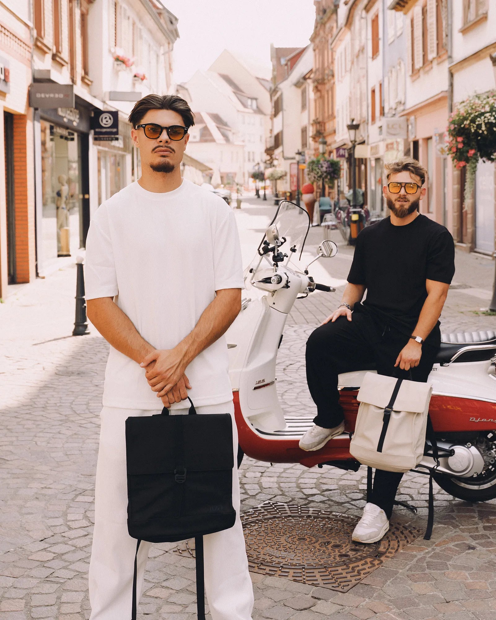 Deux hommes avec des lunettes de soleil tiennent chacun un sac à dos, l'un debout en blanc et l'autre assis sur un scooter rouge et blanc dans une rue pavée.