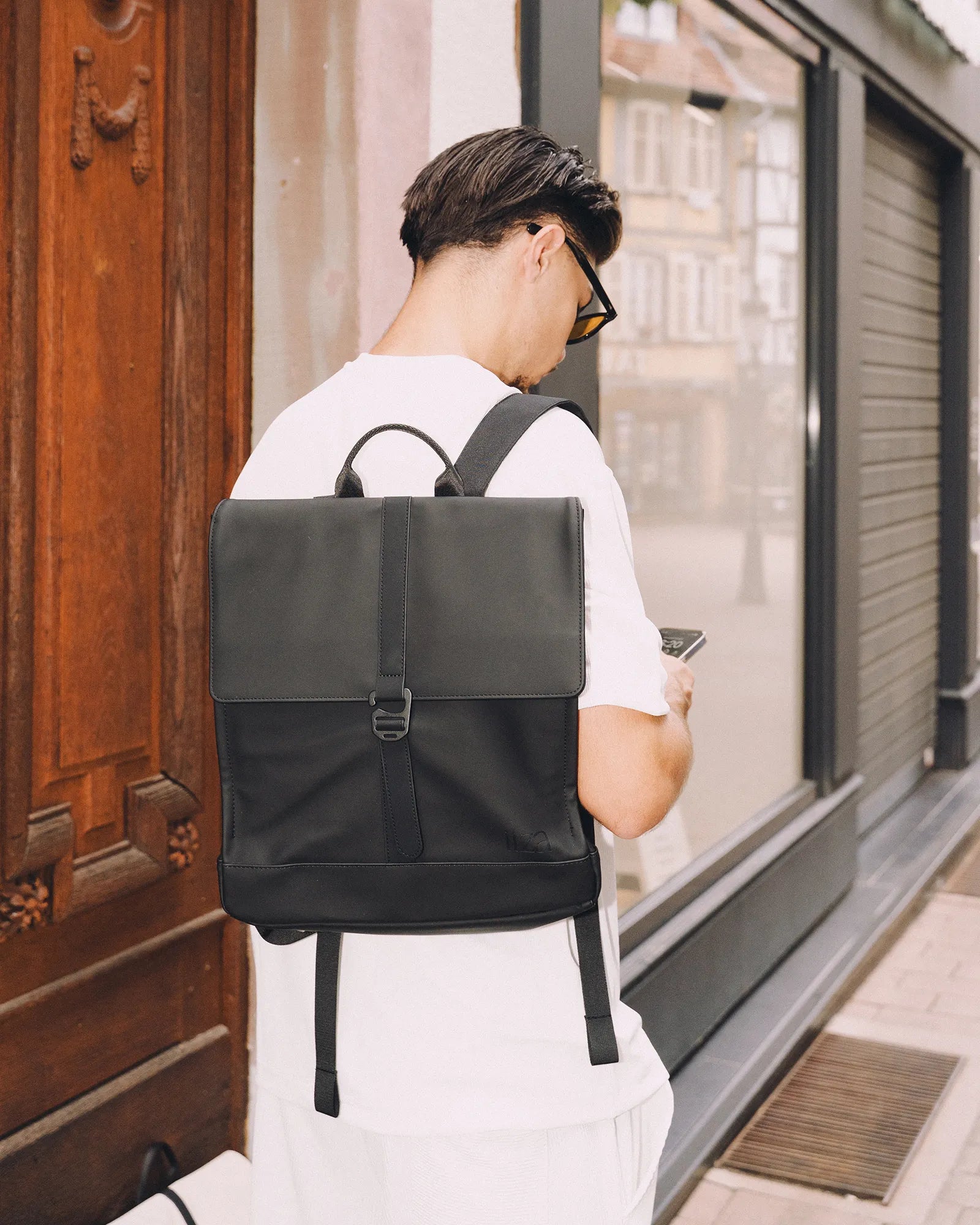 Homme portant un sac à dos noir carré et regardant son téléphone devant un bâtiment en ville.