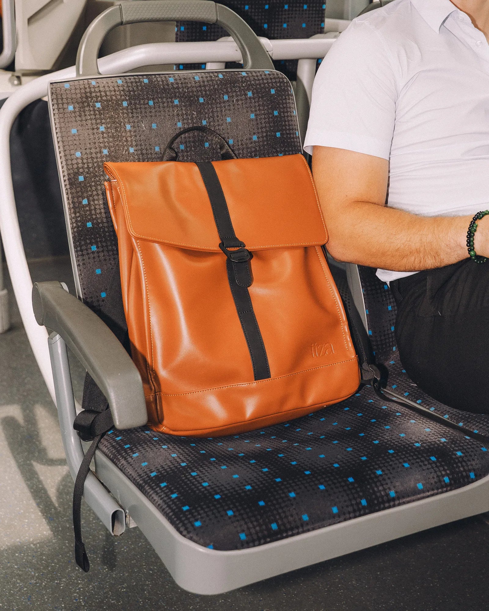 Sac à dos en cuir orange avec une bande noire posé sur un siège gris à motifs bleus dans un tramway à côté d'une personne en chemise blanche.